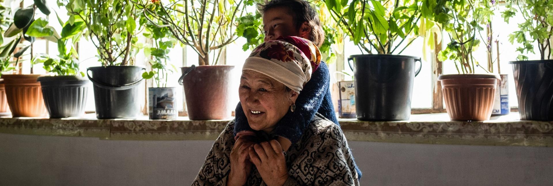 A young boy leans on a woman's shoulders, in an open kitchen-terrace setting, adorned by plants.