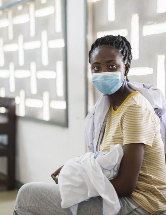 Une femme portant un masque de protection respiratoire et tenant son bébé dans les bras est assise dans une salle d'attente médicale avec d'autres femmes, le regard tourné vers l'objectif.