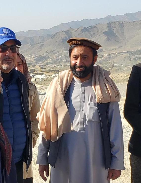 A woman in a scarf stand beside a two men in caps and a man wearing a blazer, with tan mountains in the distance.