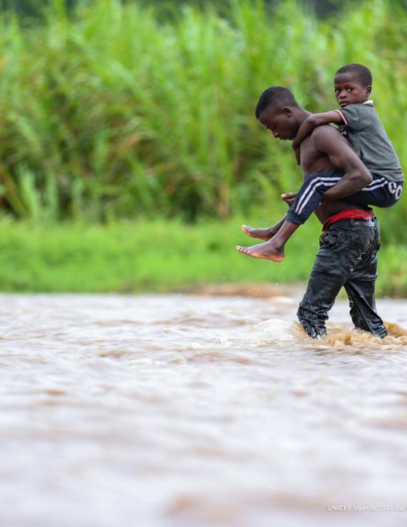 boy carries younger boy on his back as his walks through a river