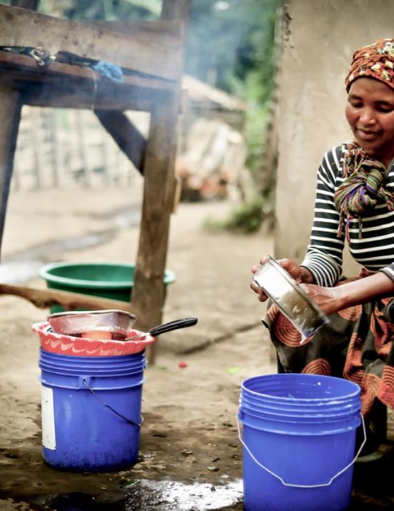 A woman sits wistfully and washes a metal pan/dish over buckets of water and soap.