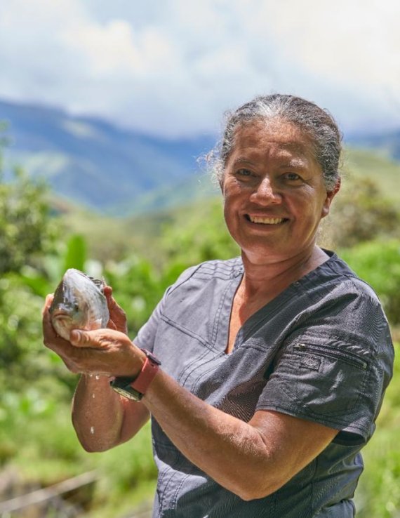 Una mujer sonriente sostiene un fruto entre las manos en un entorno rural montañoso y rodeado de vegetación.