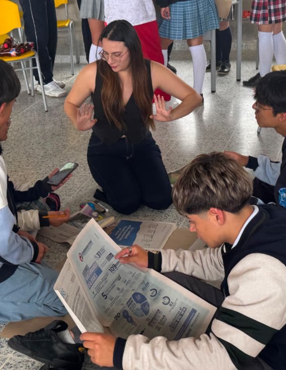 Una voluntaria dirige una actividad sobre ciudadanía digital con varios estudiantes, reunidos en el suelo con periódicos y teléfonos móviles.