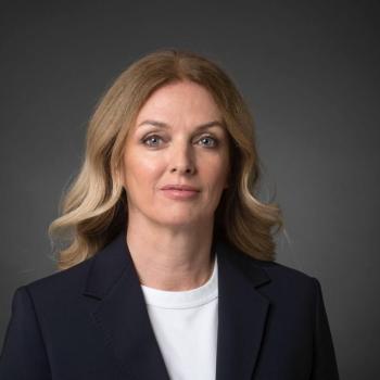 Ivana, wearing a white shirt and a black blazer, looks at the camera straight ahead.