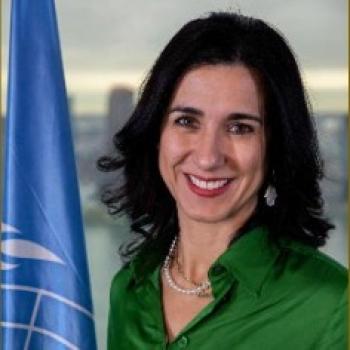 A woman in a green collared shirt stands next to a UN flag.