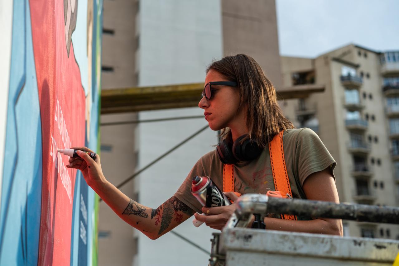 Une jeune femme aux cheveux mi-longs et portant des lunettes de soleil travaille sur une peinture murale en extérieur.