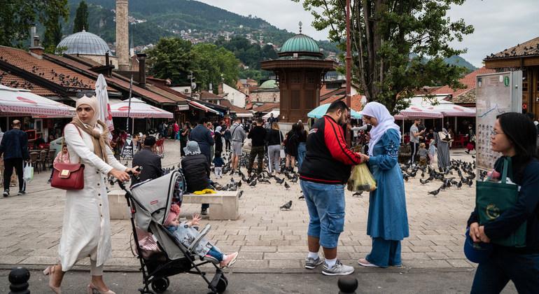 Dans une zone urbaine piétonne, au milieu d'une foule de passants, une femme portant une longue tenue blanche et un voile marche avec un enfant dans une poussette.