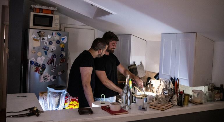 Deux hommes portant chacun un t-shirt noir sont photographiés dans une cuisine en train de discuter en préparant un repas.