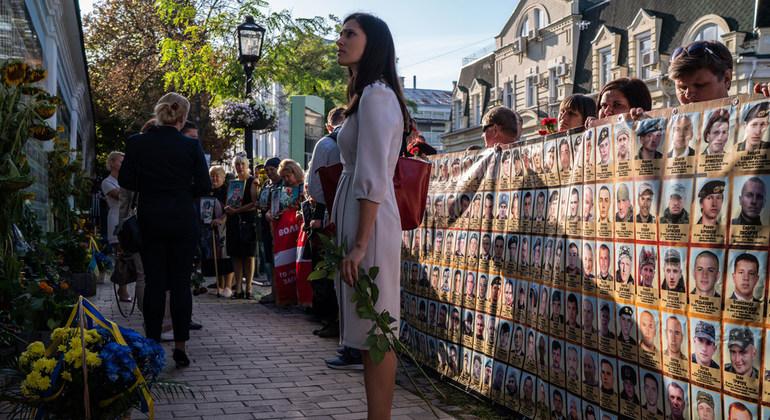 Une femme aux cheveux longs, à la silhouette longiligne et vêtue d'une robe blanche se tient debout dans une rue piétonne, une rose à la main, près d'un cordon de manifestants tenant une grande banderole portant des portraits-photos.