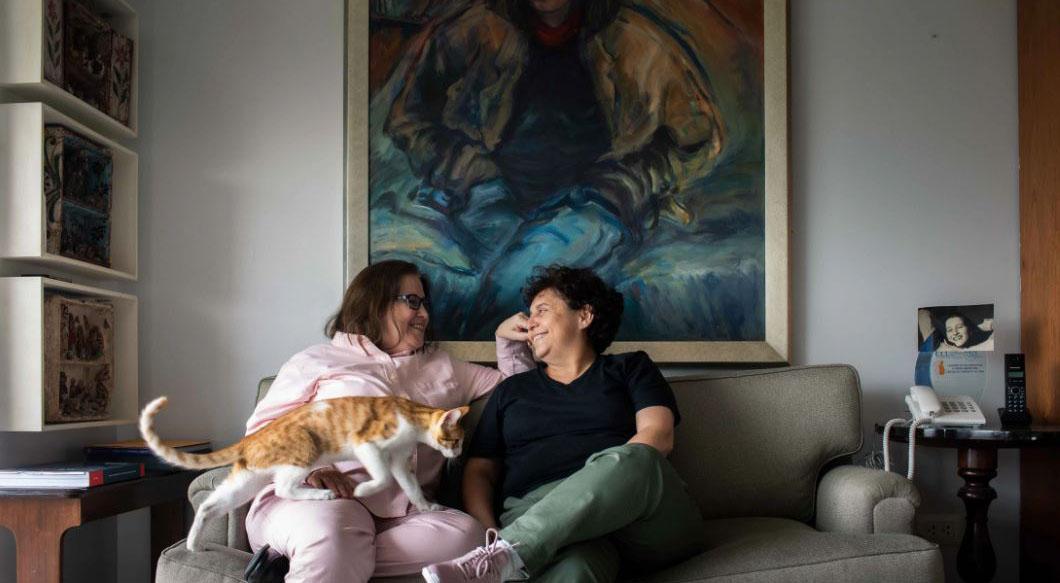 Susel and Gracia lovingly look at each other and smile, while sitting on a couch with a cat making its way to their lap.