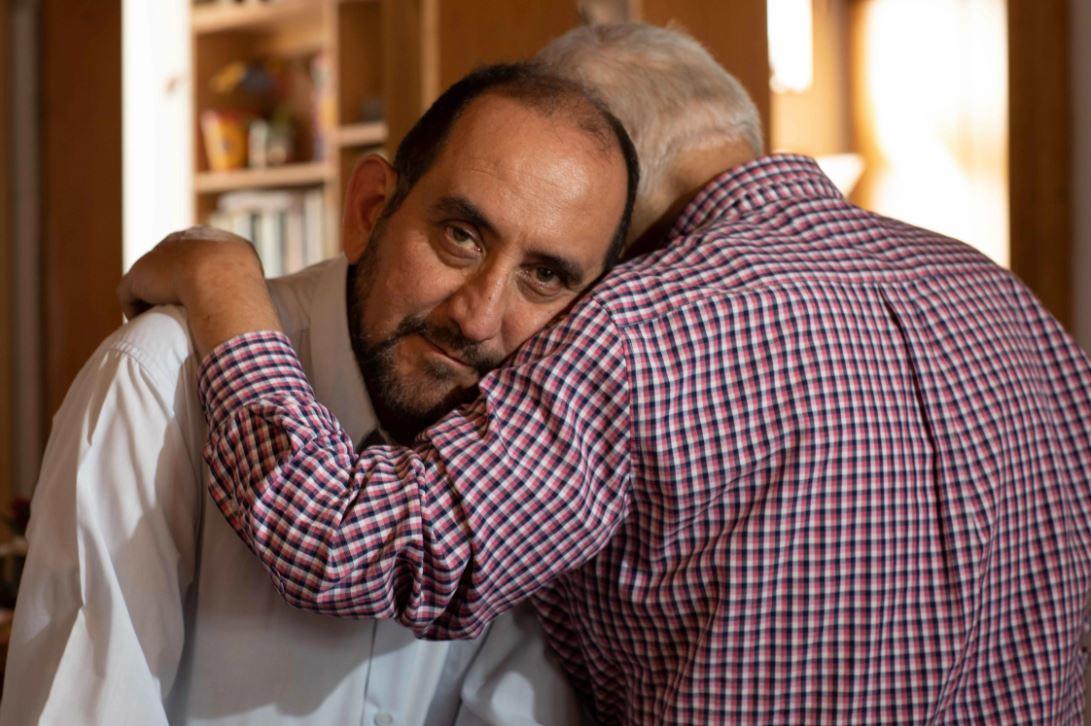 Alejandro faces the camera as Camilo sits opposite him with his back to the camera and his arm lovingly draped around Alejandro. 