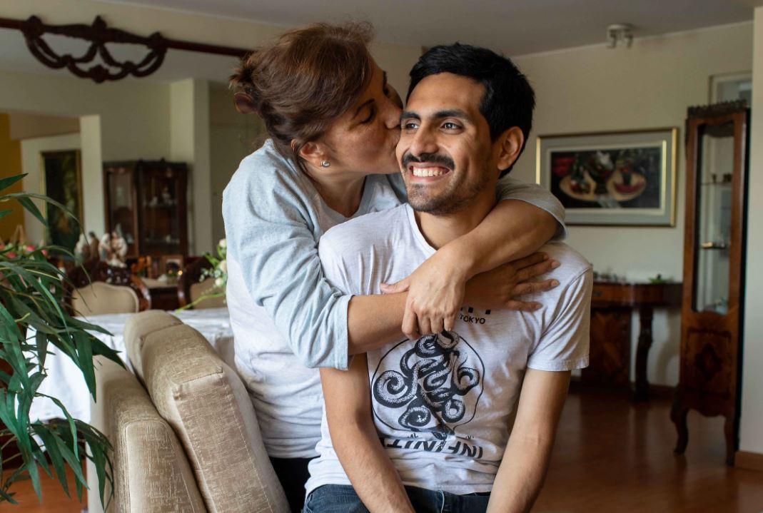 Cecilia, mother, wraps her arms around her son, Carlos, while she kisses his cheek and he smiles. 