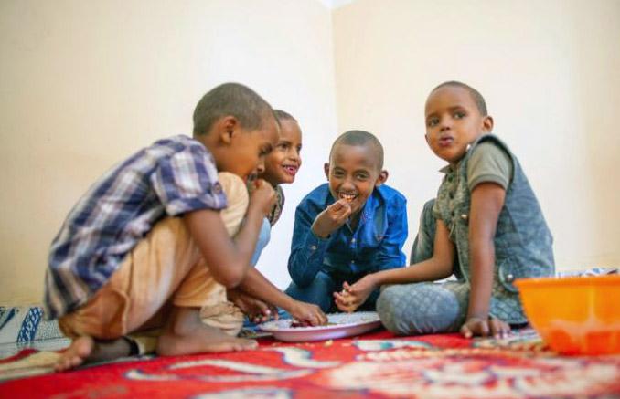Shows a photo from a low angle of the boys sitting around a plate of food, eating and smiling at the camera and each other. 