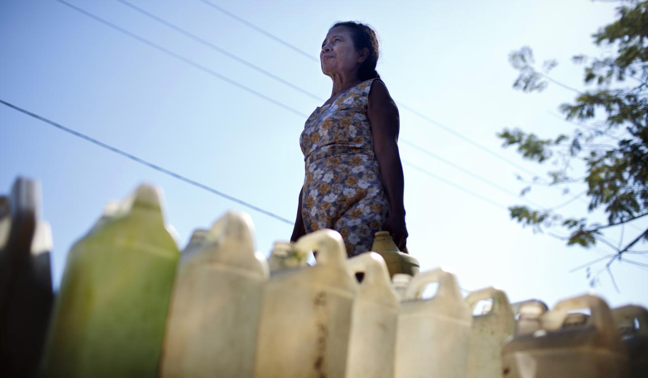 La mujer con gesto de determinación camina con un bidón de agua. En primer plano hay una fila de bidones de agua sobre el suelo.