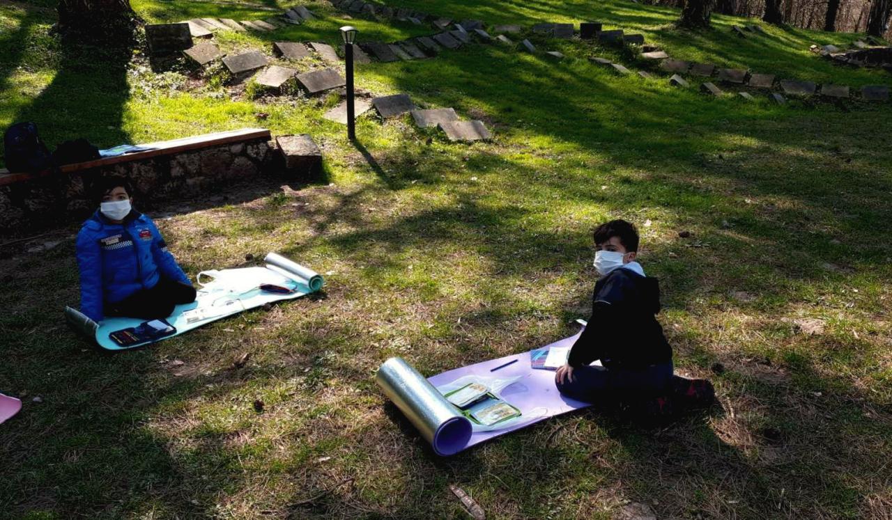 Young students wearing protective gear take their online courses outside.