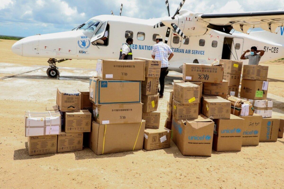 Un avión con el emblema del PMA aparece en el fondo, con varios montículos de cajas de suministros en primer plano.