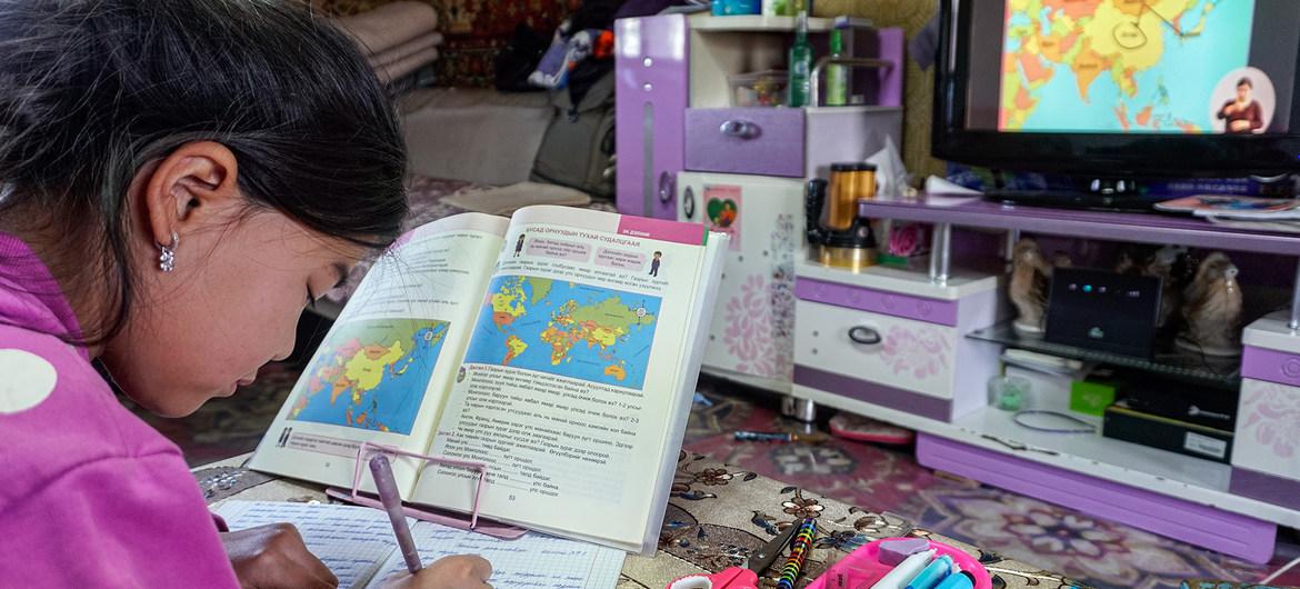 Une petite fille fait consciencieusement ses devoirs, assise à une table où sont disposés un livre de géographie et des jouets.