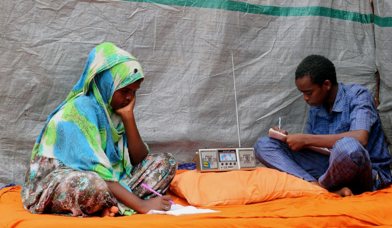Two young people sit by a portable radio and work on school assignments.