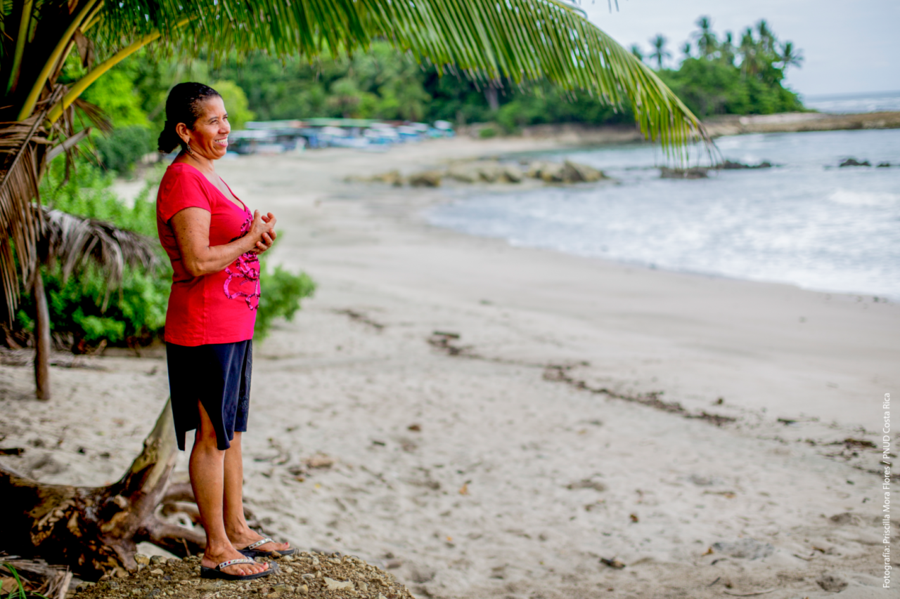Woman smiles as she pensively as she stands on a beach looking towards the ocean. 