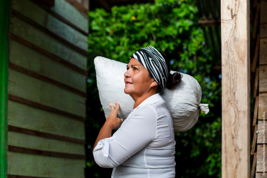 A woman looks up stoically as she holds a large bag over her shoulders.