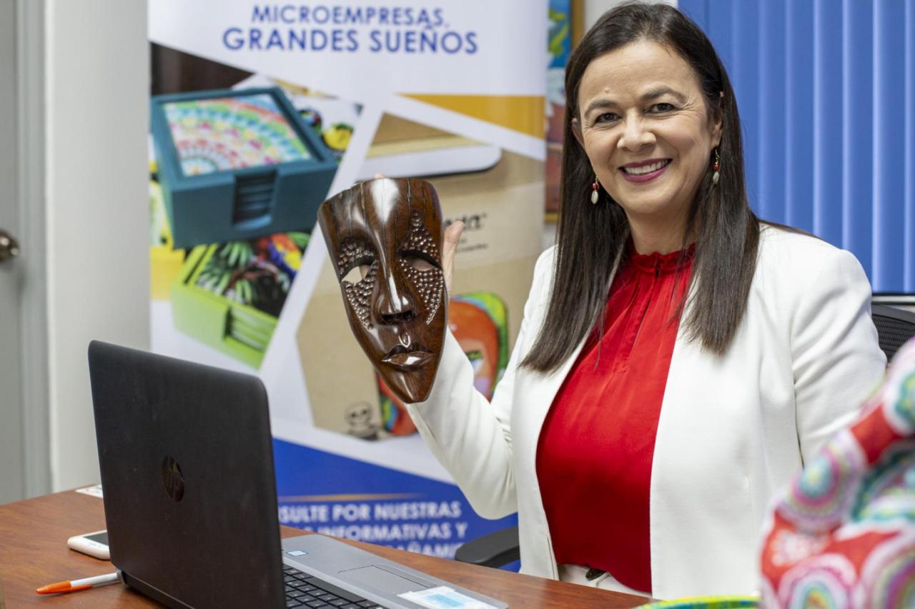 A woman smiles happily as she stands at a desk holding a mask.