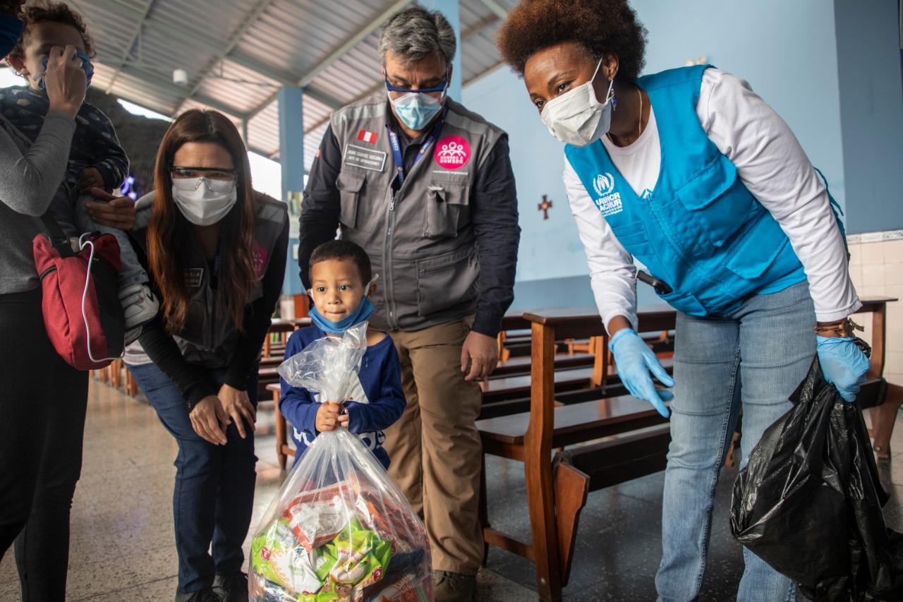 Personal de la ONU y de ONGs acompañan a un niño mientras sostiene una gran bolsa de alimentos y suministros donados.