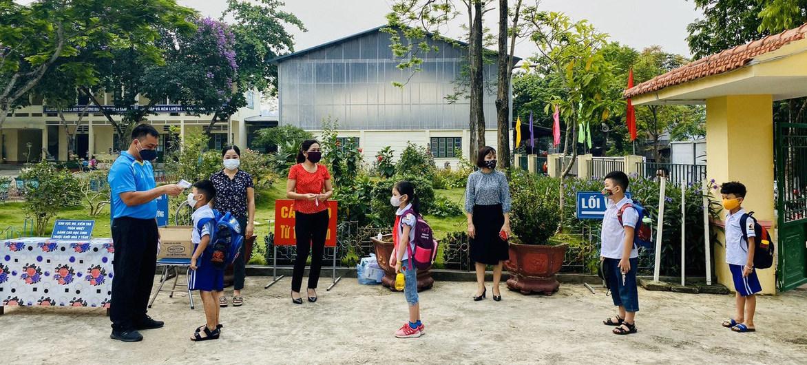 Dans l'enceinte d'une école, de jeunes élèves portant des masques de protection et encadrés par des membres du personnel enseignant font la queue en se présentant pour un contrôle de température.