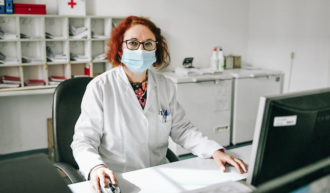 Snežana Bursać Aranđelović está sentada en su escritorio con una computadora (frente a ella) en el Instituto de Salud Pública del Cantón de Sarajevo. Lleva una bata blanca y una mascarilla quirúrgica azul y está mirando a la cámara. Podemos ver detrás archivos y mobiliario de oficina colocado en estanterías blancas.