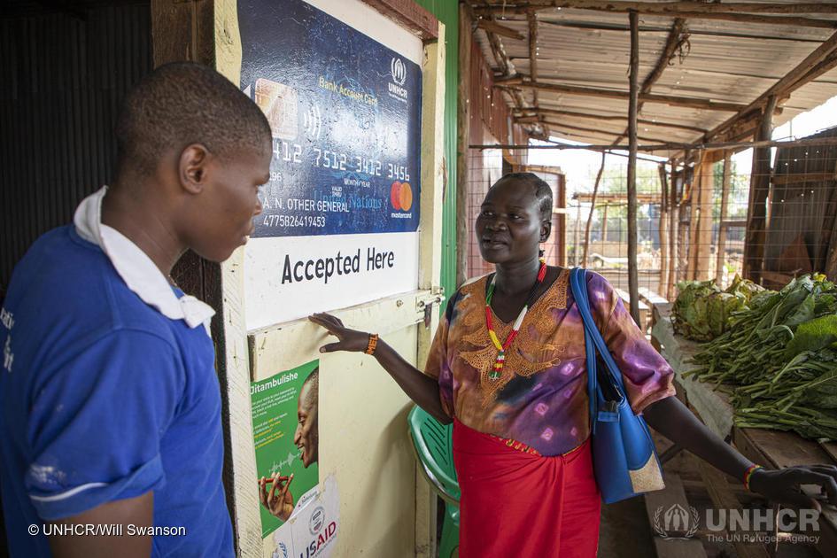 A woman stands by a walk covered in signage while speaking with a man, which stands by the doorway of the market at the Kalobeyei Settlement.