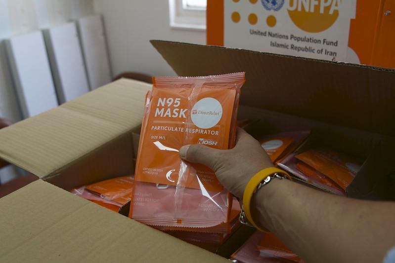 A hand holds up packaged PPE above a UNFPA-donated box.