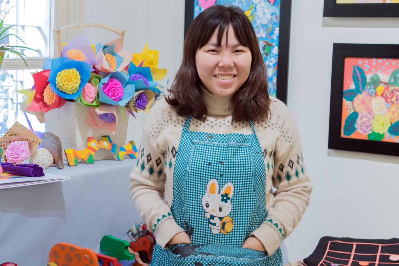 Tu Thanh Thuy sonríe alegremente a la cámara mientras está de pie con las manos en los bolsillos de su delantal frente a coloridas flores de papel y pinturas.