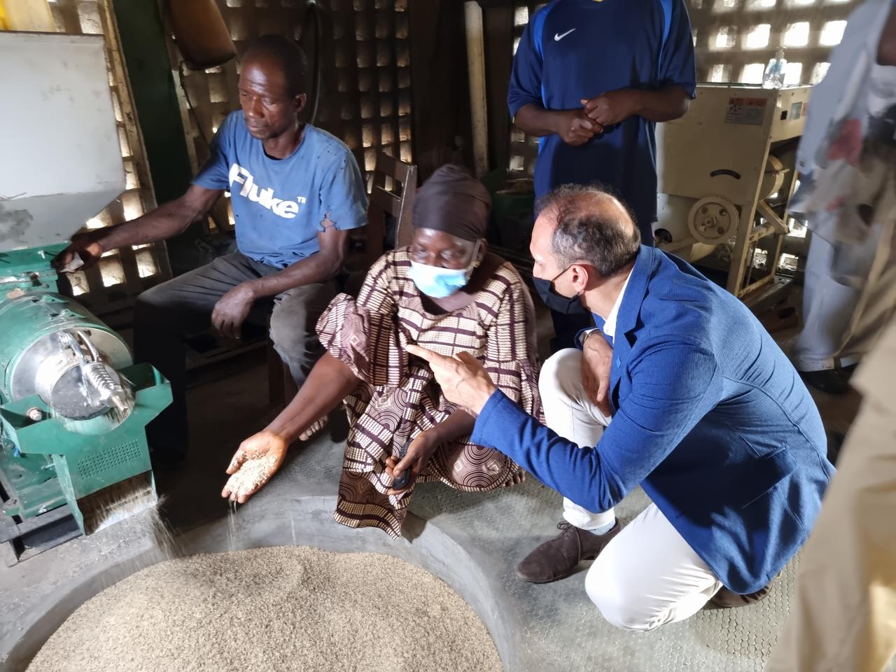 Resident Coordinator, Vincent Martin kneels by a woman community leader as they speak near a machine that is filling a round hole with grains.