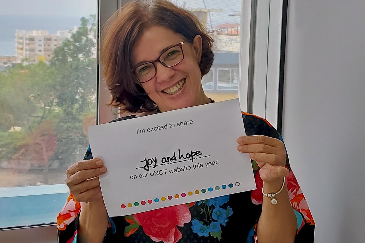 Anita warmly smiles as she holds a sign that reads she's excited to share joy and hope on their country team website.