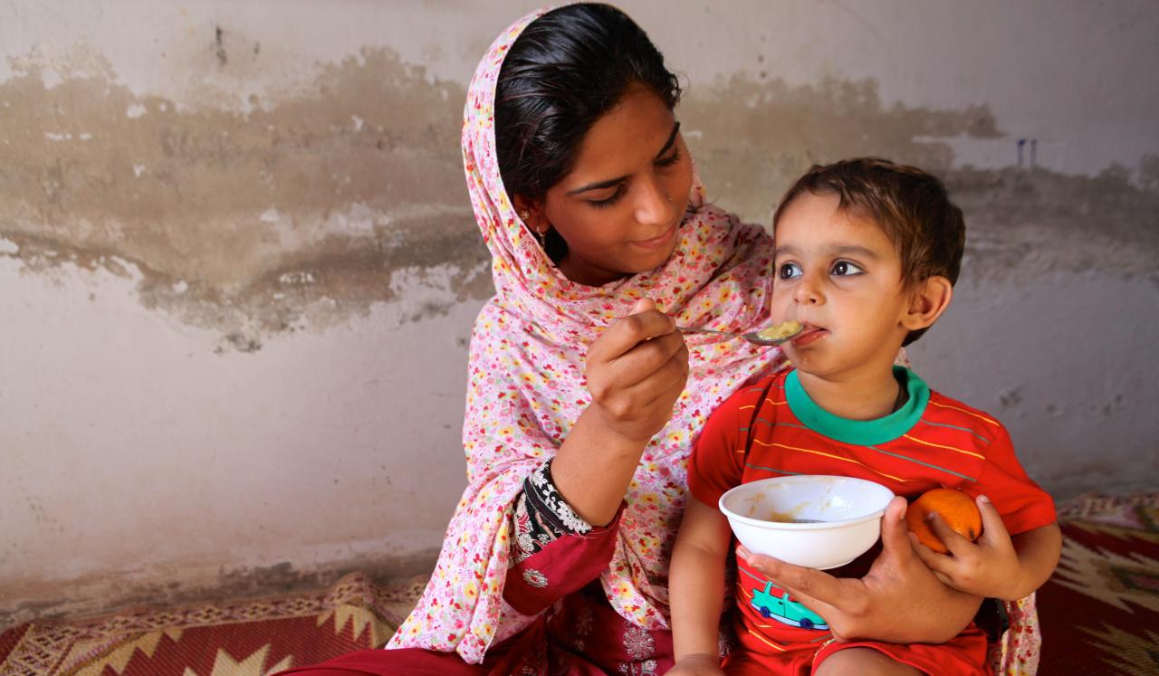 A young woman holds a child in her lap while she feeds him from a bowl.