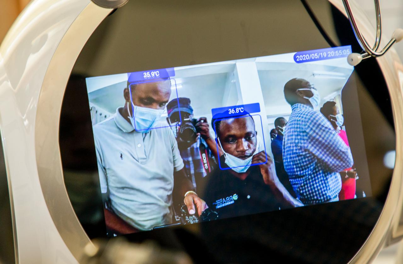 The screen of one of the robots shows the faces of two gentlemen with their body temperatures noted on the screens above their heads.