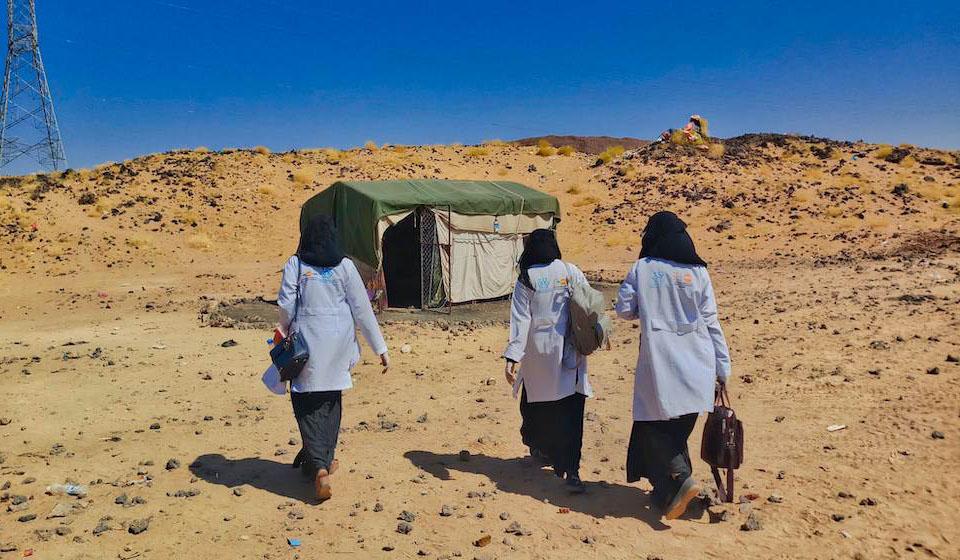 Tres parteras con batas de laboratorio se alejan de la cámara caminando, en dirección a clínica de salud móvil.