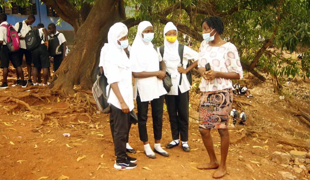 Una mujer con una mascarilla facial está de pie y habla con tres adolescentes.