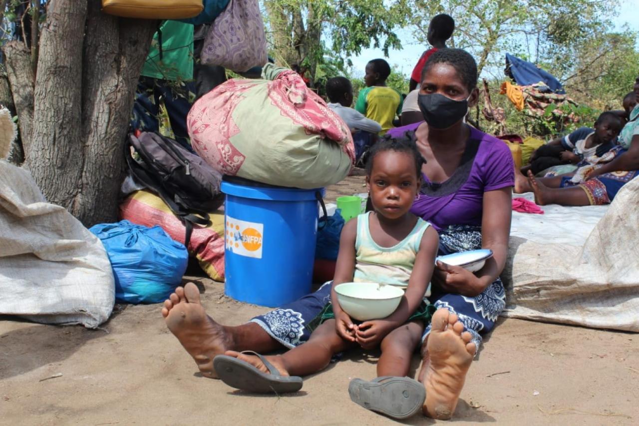 A mother and child sit on the ground together holding bowls for food with the possessions to the left of them, including a dignity kit by UNFPA.