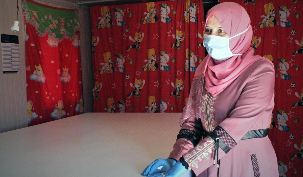 Haifa, 33, wears a face mask and gloves as she stands by a table in a room with red curtains.