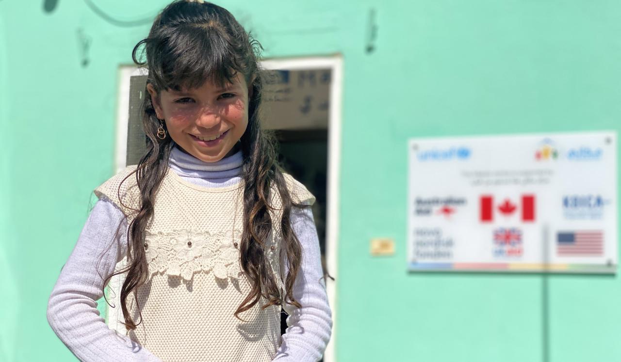 Kawthar, 10, smiles shyly at the camera as she stands outside in front of a green building.