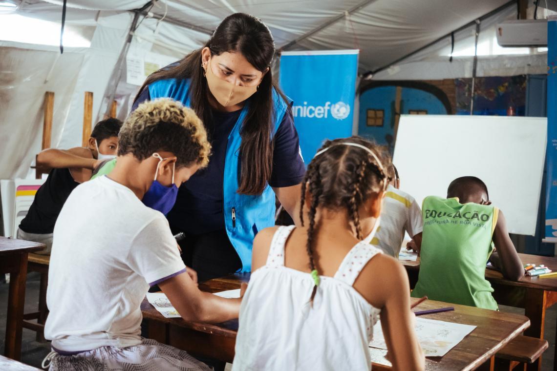 A teacher with a face mask and blue vest talks to a boy and a girl in a classroom. 