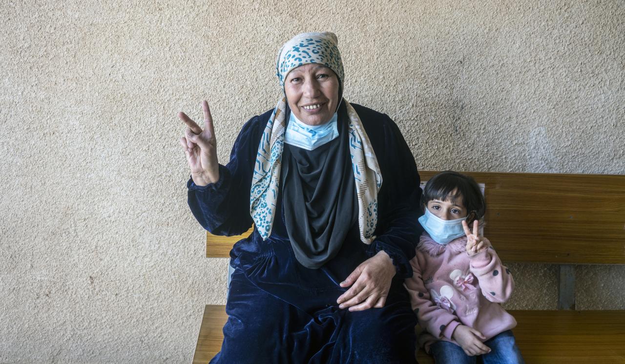 A woman smiles cheerfully at the camera with her young daughter. Both flash the 'V' for vaccine sign with their fingers.