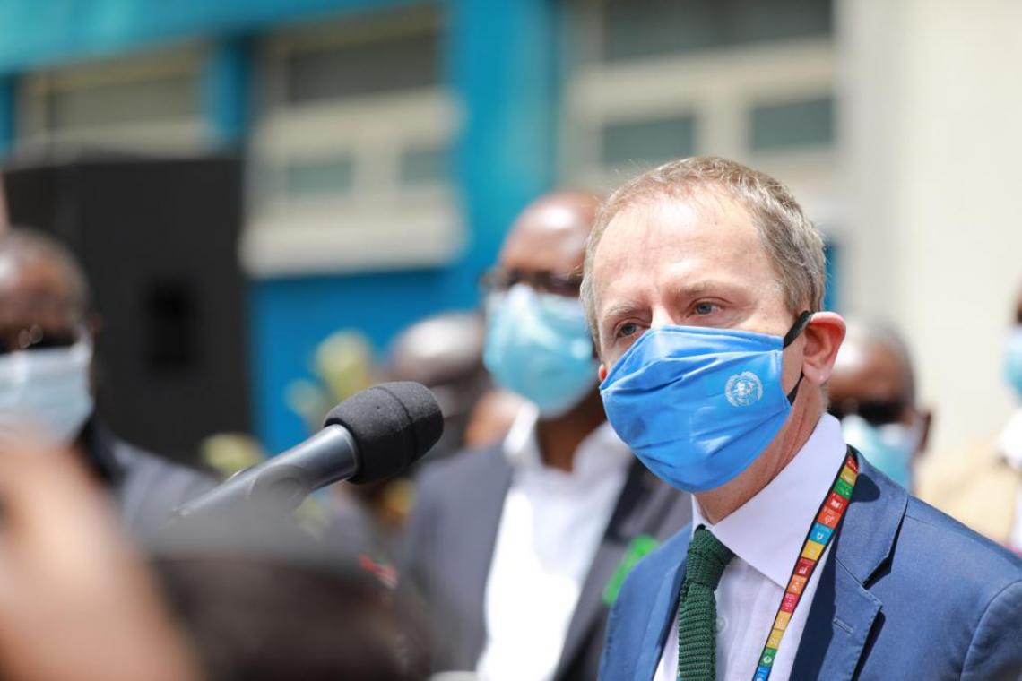 Personas con máscaras están detrás de un hombre, con una máscara azul claro, que usa una corbata verde y un traje azul. El hombre se encuentra hablando por un micrófono.