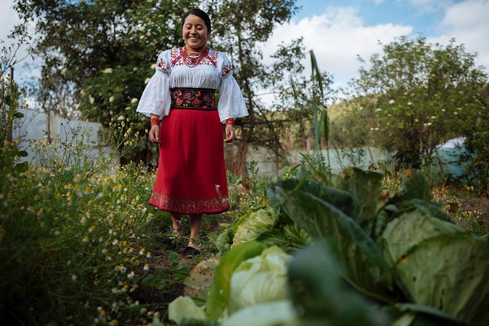 Una niña sonriente con atuendo tradicional se encuentra en un campo con árboles y flores verdes.