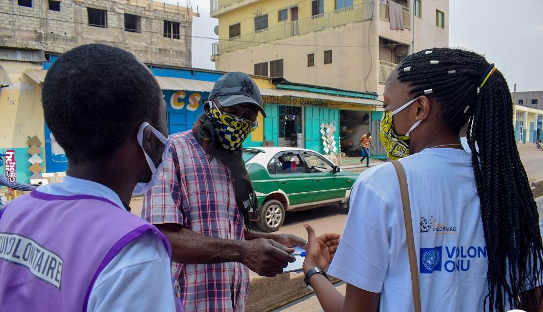 Three people in masks stand on the side of the road as a green car passes by behind them.