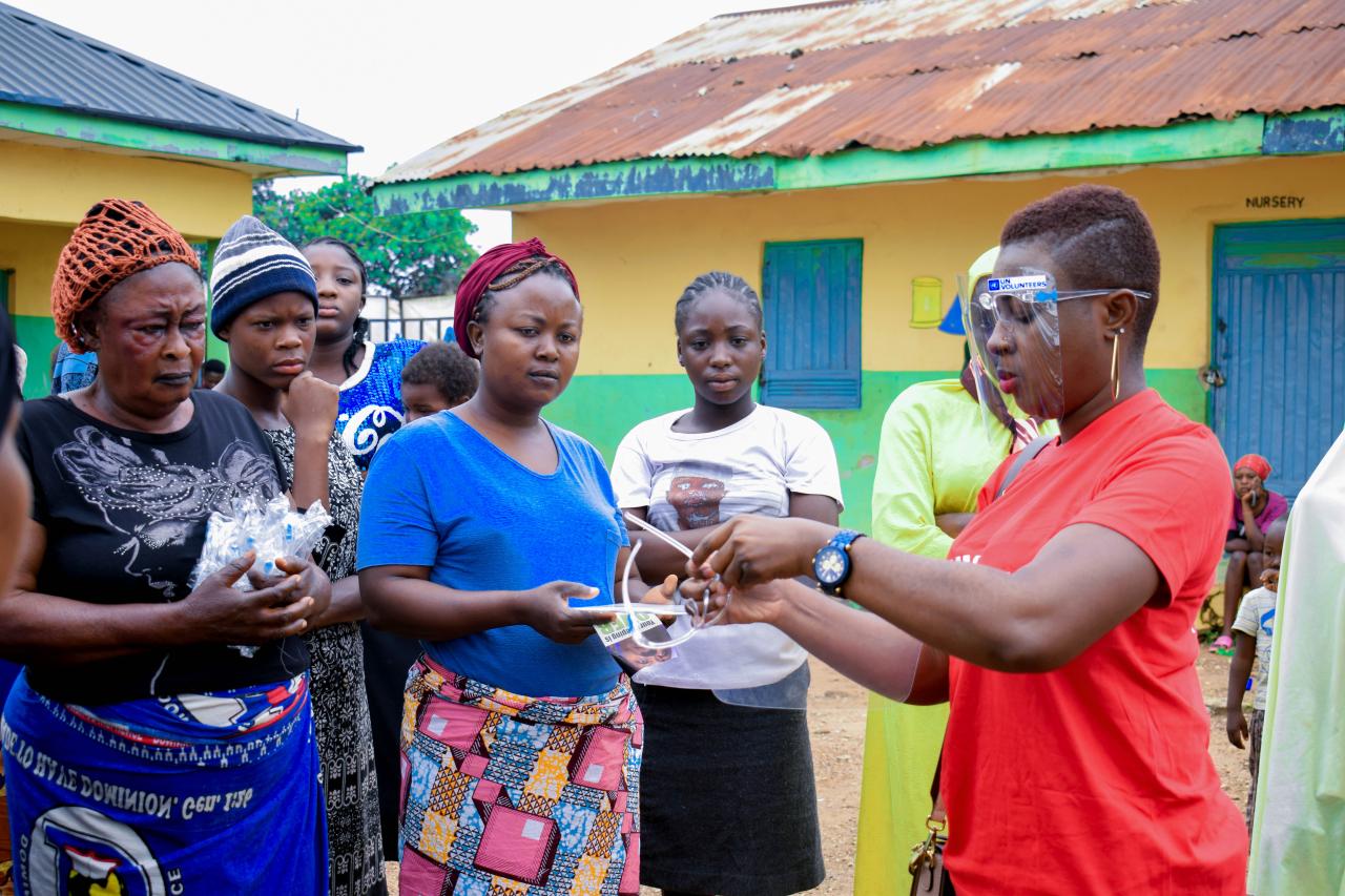 A woman in a red shirt and face shield shows a group of women and girls a pair of goggles.