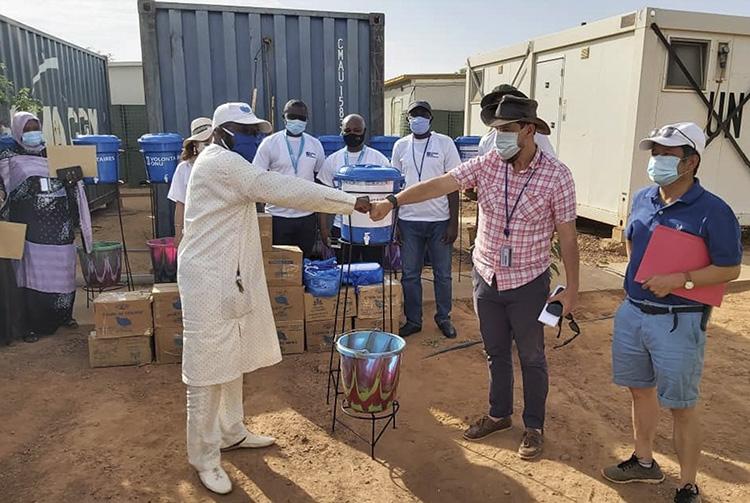 Two men fist bump their hands while several other people stand around them near large containers.