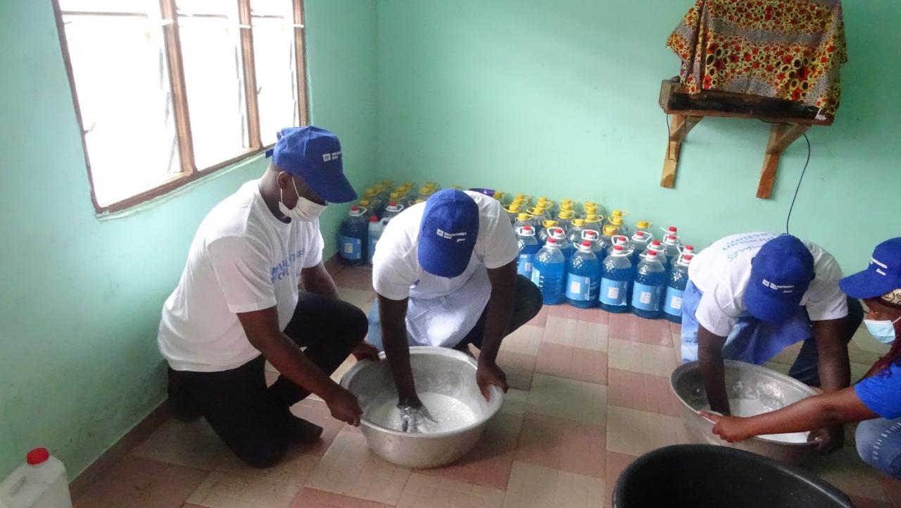 Four people kneel on the ground with two metal pails. Two individuals have their hands in the pails and are mixing up a white substance.