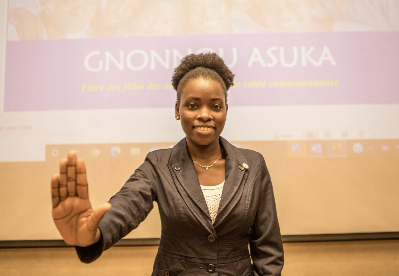 A young woman in a white shirt and black blazer extends her palm at the camera.