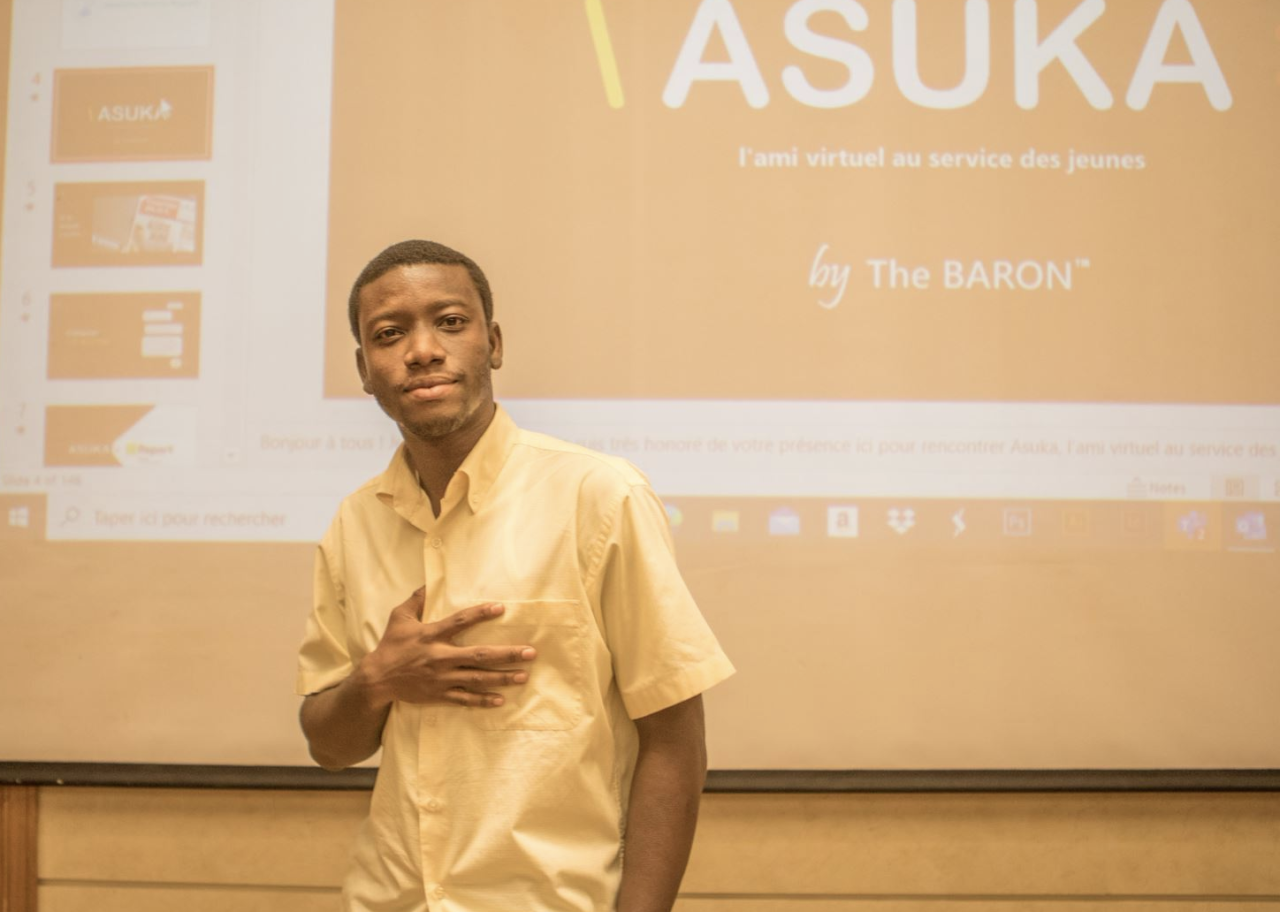 A young man, in a white button down shirt, stands in front of a projector holdings his hand to his heart. 